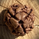 Top view of 3 Mukhi Rudraksha placed on a wooden block with a blurred background, showing its natural surface and three-lined structure.